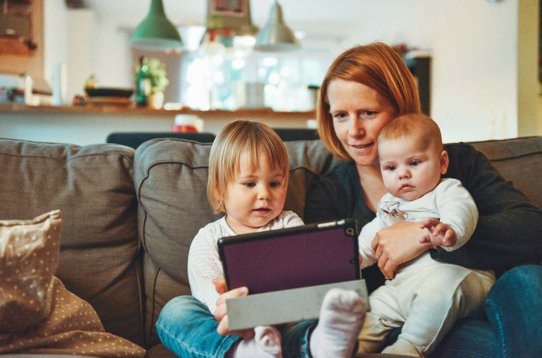 Mother in her home holding two small children on her lap while they all look at iPad.